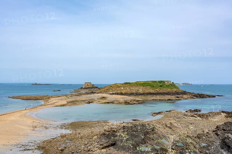 A picturesque view of the uninhabited tidal island of Grand Bé at low tide in the Bay of Saint-Malo. The historic Fort du Petit Bé can be seen on the left of the horizon. Photographed from the historic city walls (Remparts) on a clear day with a blue sky above the rocky Atlantic coast in the province of Ille-et-Vilaine, Brittany, France
