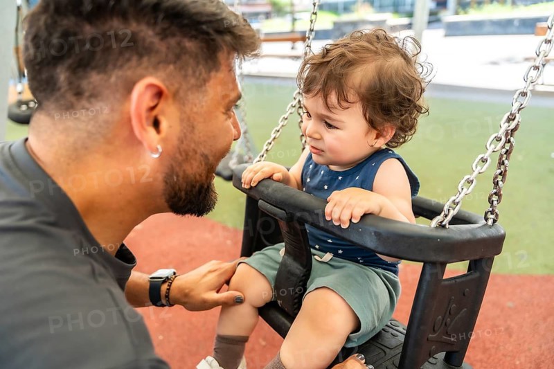 Father kneeling and comforting his sad toddler son sitting on a swing at the playground, creating a touching moment of paternal care and support during playtime outdoors
