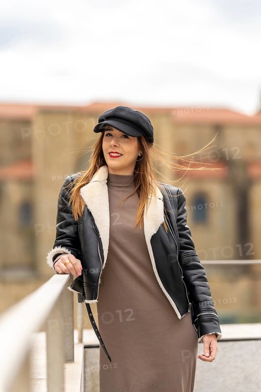 Portrait of a fashionable woman posing in a city environment, wearing a black cap, leather jacket, and a stylish dress