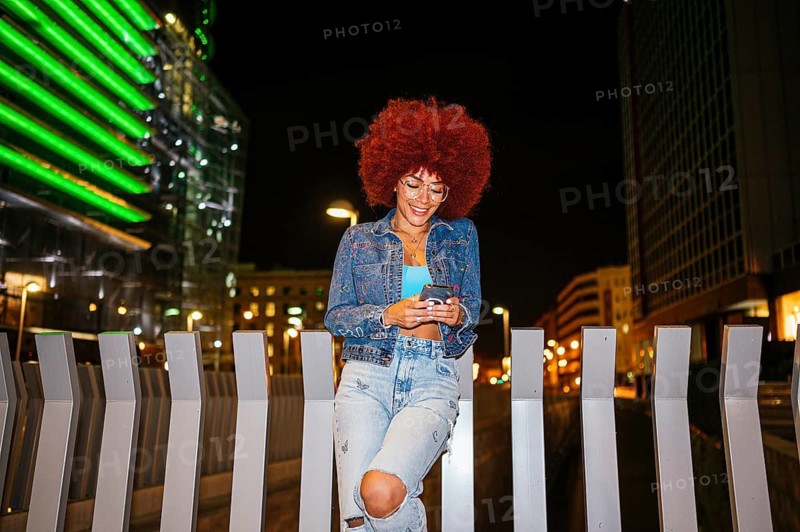 African descendent woman with vibrant afro hair smiles while using her mobile phone, casually standing in an urban setting at night, illuminated by city lights