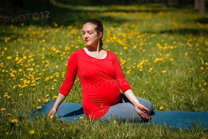 Pregnancy yoga exercise, pregnant woman doing asana Marichyasana easy variation outdoors on grass lawn with dandelions in summer