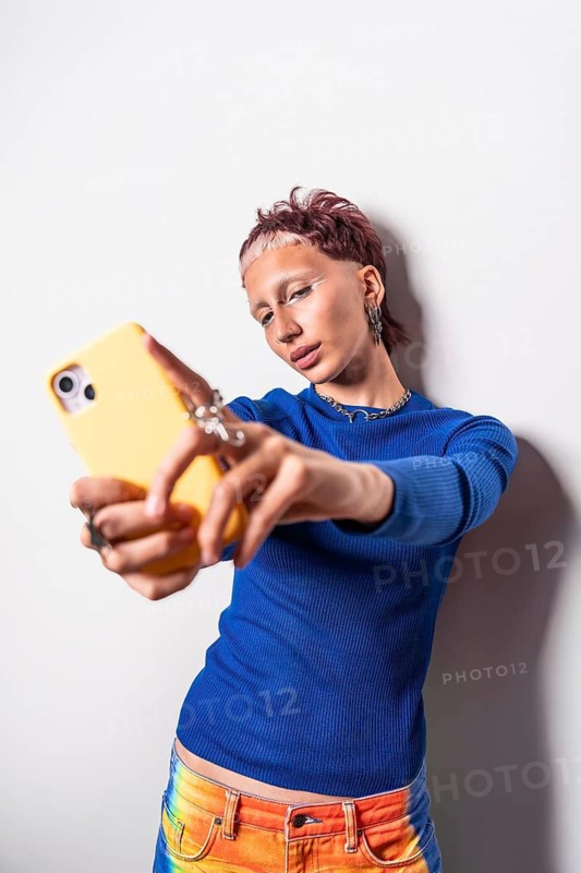 A trendy woman wearing colorful trousers and a blue top takes a selfie with her yellow phone, showcasing modern fashion and vibrant style against a white backdrop