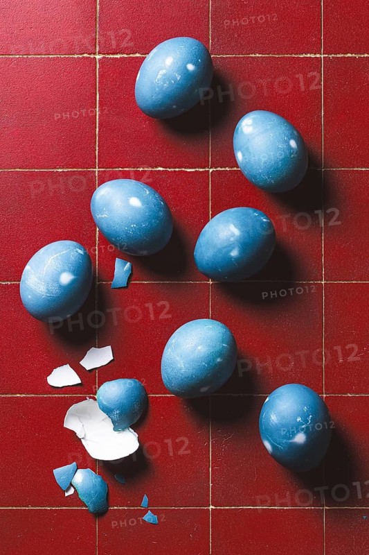 From above, a collection of blue Easter eggs on a red tiled floor, some whole and one broken with scattered pieces