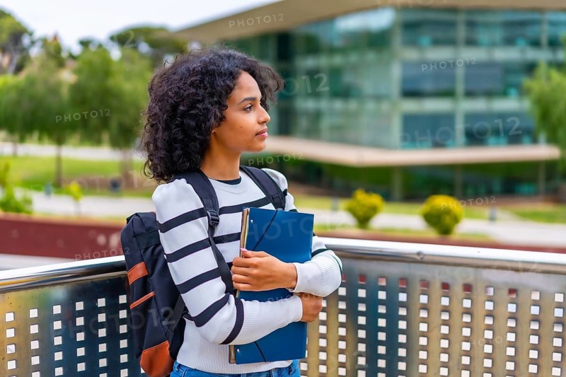Side view of a serious latin woman standing holding a folder outside the university campus