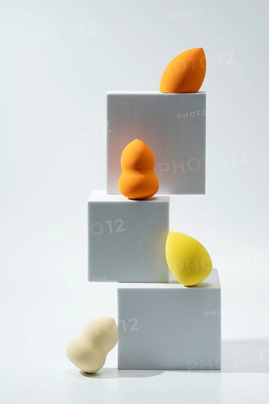 A set of vibrant cosmetic sponges in various shapes and hues is creatively displayed on stacked white geometric blocks The minimalist background adds elegance and focus