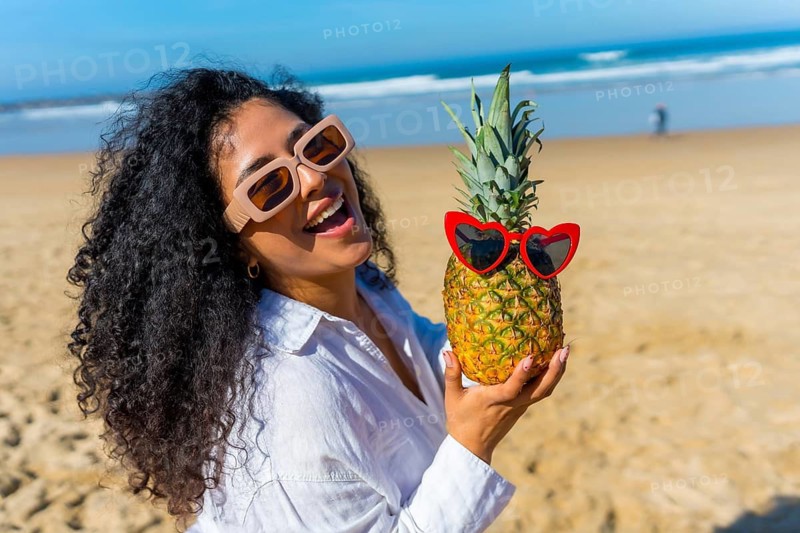 Young woman with curly hair wearing sunglasses and a white shirt, laughing while holding a pineapple with heart shaped sunglasses, enjoying a tropical summer vacation by the ocean