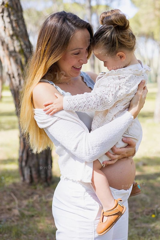 A pregnant woman joyfully embraces her young daughter in a serene outdoor setting. The scene captures a tender moment of mother-daughter connection and familial love