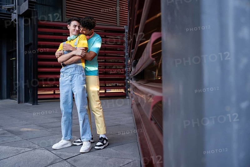 A gay couple share a tender embrace outside an urban building. They are dressed casually, exuding happiness and connection in the city setting
