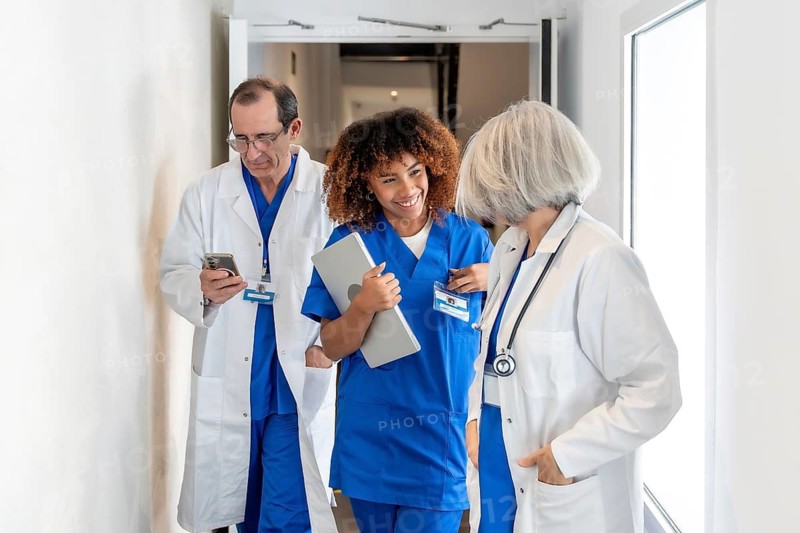 Diverse healthcare team walking a bright hospital corridor, young african american nurse with laptop smiling and talking to a senior female doctor while a male doctor checks his smartphone