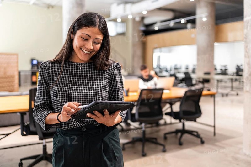 A woman in a modern office smiles as she uses a tablet, with colleagues in the background The image conveys teamwork, collaboration, and technology in a professional setting