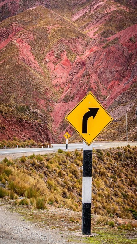 A road sign indicating a curve stands before stunning red-striped mountains. Captured in the Peruvian Andes, the scene highlights the region's unique geology and beauty