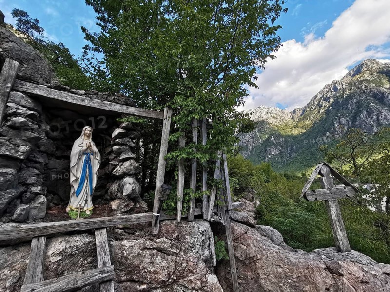 A Jesus statue in a small stone grotto with a wooden cross with a view of the surrounding mountains, peaceful nature, peaks of the Balkans, idyllic landscape, hiking in the Albanian Alps National Park