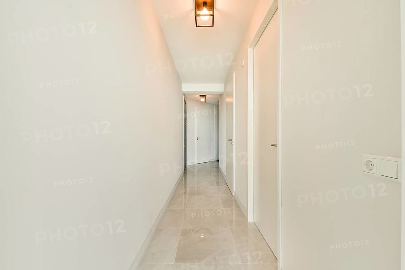 A bright, minimalist hallway with white walls and doors, featuring sleek tiles and modern pendant lights casting a warm glow, creating an inviting atmosphere