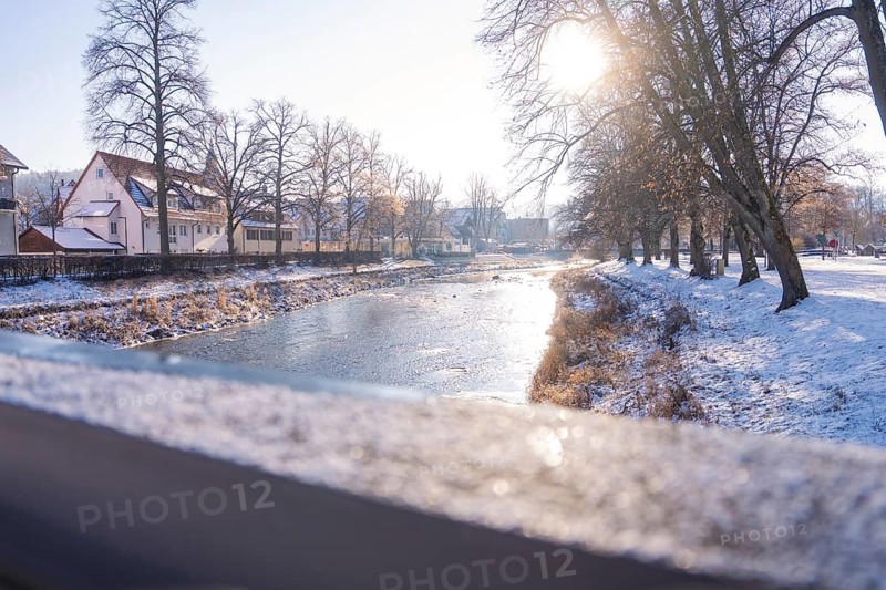 Snowy riverbank in winter with sunlight and surrounding houses, Nagold, Calw district, Germany