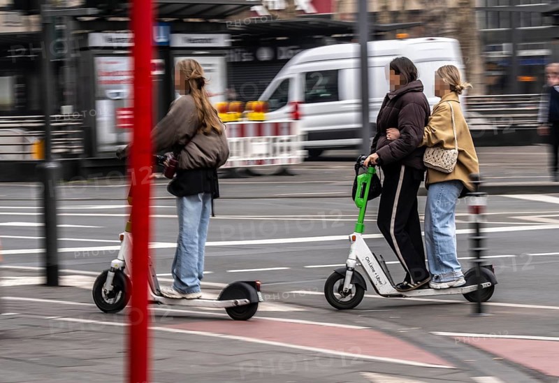 Young woman, two of them on an e-scooter, in the city centre, here at Neumarkt in Cologne, parked bikes and e-scooters at the roadside, bicycle lane, Cologne, North Rhine-Westphalia, Germany