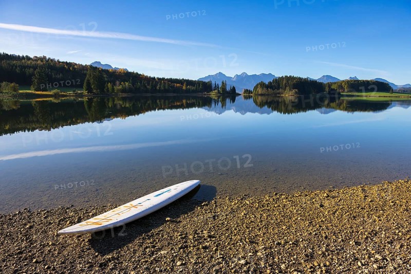 A calm lake with a surfboard on the shore, surrounded by mountains and a clear blue sky, Forggensee near Füssen, Ostallgäu, Bavaria, Germany