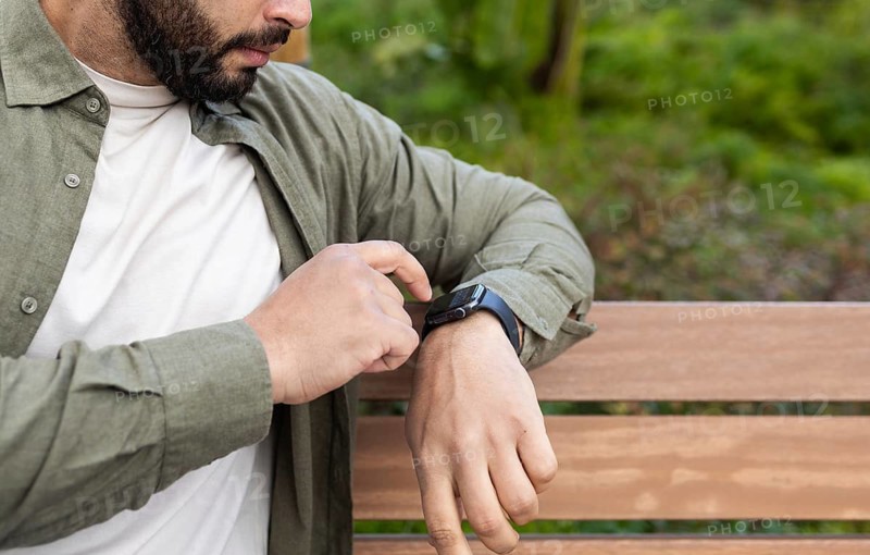 A man interacts with his smartwatch while sitting on a wooden bench in a lush in the city. He wears a casual green shirt over a white tee, focusing on his wrist device