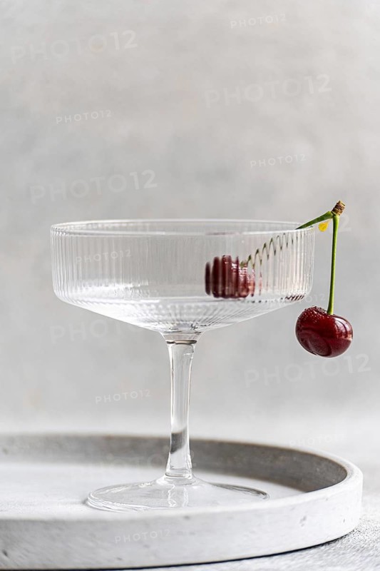 A ripe cherry delicately dangles off the edge of a clear glass, set against a textured concrete background, creating an artistic and sophisticated visual contrast