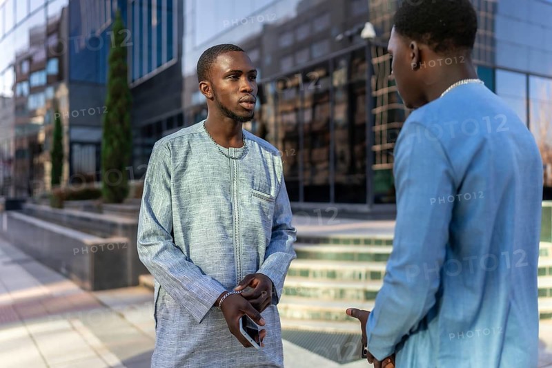 Two senegalese businessmen wearing traditional dashiki attire are having a conversation outside modern office buildings