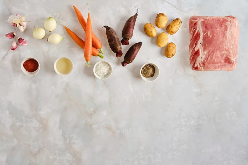 Fresh vegetables and herbs on countertop. Carrots, potatoes, beetroot and onions sit next to pieces of meat. Spices and oil are ready for cooking. This moment shows meal preparation in kitchen