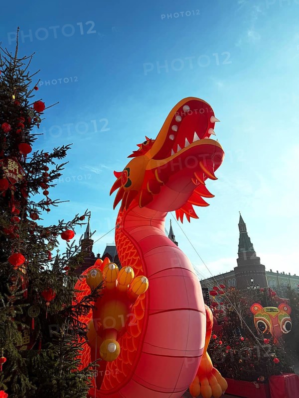 A giant red dragon figure stands tall among decorated trees during a Chinese New Year celebration, symbolizing good fortune and strength, with festive elements all around