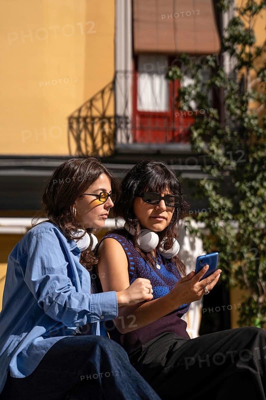 A multiethnic lesbian couple enjoys a sunny day outdoors, wearing headphones and sunglasses, exemplifying diversity and LGBTQIA+ pride in a relaxed urban setting
