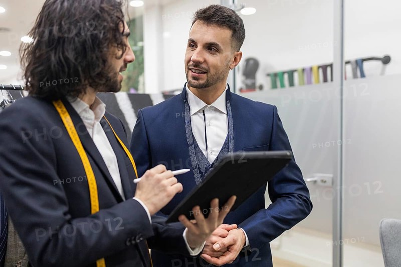 A tailor with a measuring tape consults with a client holding a tablet in a suit store. The interaction highlights professional tailoring services