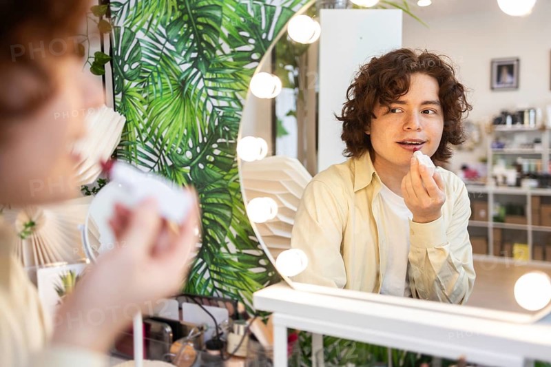 A young man applies lipstick while looking into a mirror surrounded by lights in an organic beauty shop The eco-friendly store features natural cosmetics and lush green decor