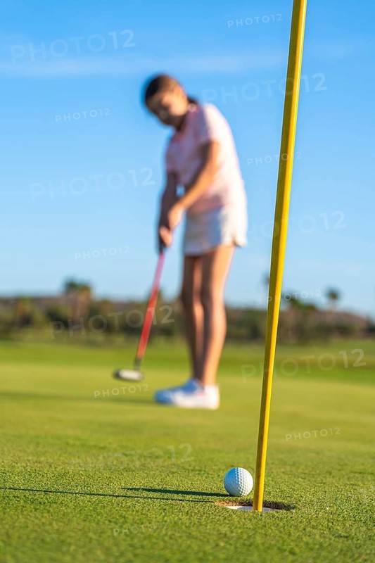 Vertical photo with focus on golf hole while girl playing on the background