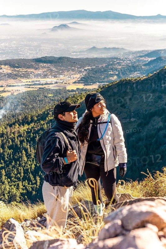 Two friends stand on a mountain overlooking a vast valley, enjoying the scenic view Dressed in winter gear, they embrace the adventure and camaraderie of the hike