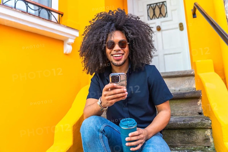 Young man with curly hair and sunglasses using mobile phone and holding reusable coffee cup while sitting on steps in front of a vibrant yellow building