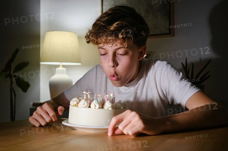 A teenager in a white shirt blows out the candles on a birthday cake in a dimly lit room. The scene captures a moment of celebration and joy