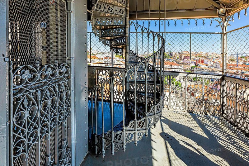 Ornate wrought iron balcony overlooking a sunny cityscape with clear blue sky, Elevador De Santa Justa, Lisbon