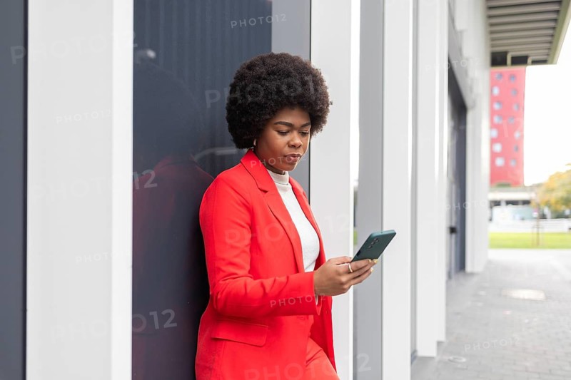 Confident woman in a vibrant red suit leans against a modern building, engaged with her smartphone. The urban backdrop and striking attire highlight her professional allure