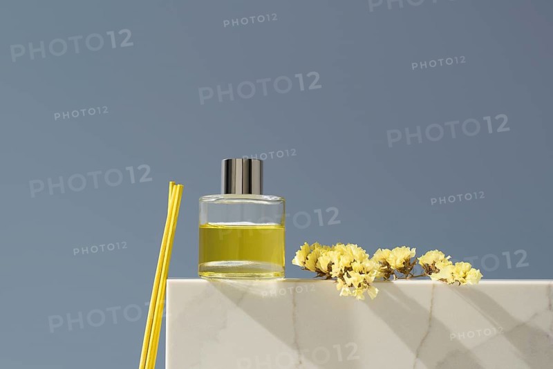 Elegant glass diffuser filled with lemon-scented liquid, accompanied by yellow sticks and delicate flowers, against a minimalist background. Fresh and decorative
