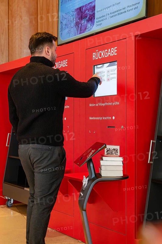 A man interacts with a return machine while a robot carries books, Interactive guided tours by robots Personalities of the company Robolutions, Pforzheim, Germany