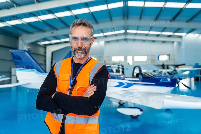 Mature bearded aviation technician in orange safety vest and protective glasses stands with arms crossed in an aircraft hangar by small planes, confident and professional