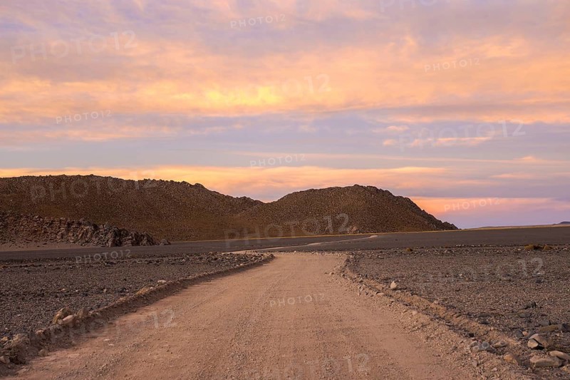 A serene sunset casts warm hues over a scenic gravel road winding through the rugged terrains of La Puna in Argentina, illustrating a peaceful yet unyielding landscape