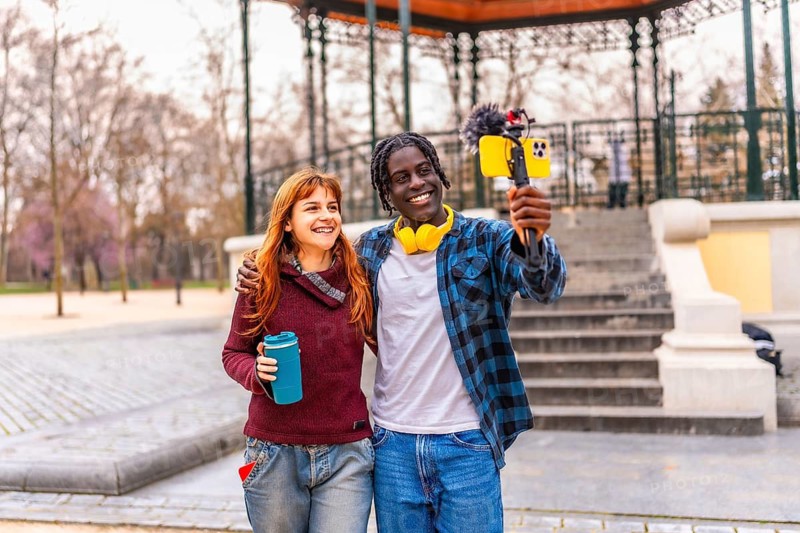 Two young content creators are recording a vlog in a park, holding a smartphone with a microphone attached