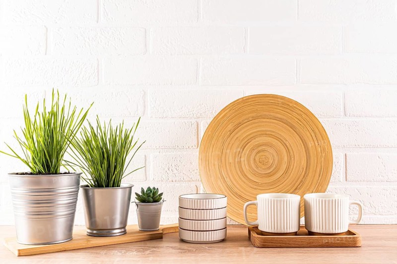A contemporary kitchen setting featuring a wooden round plate, white striped mugs on a tray, and various potted green plants against a white brick wall