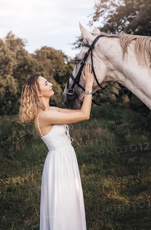 A young woman in a flowing white dress gently strokes a white horse in a picturesque outdoor setting, highlighting the bond between them. Trees and soft sunlight add to the serene mood