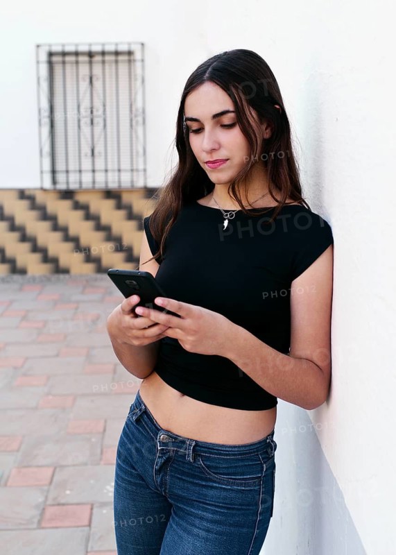 A young woman in casual jeans and a black top leans against a white wall, focused on her smartphone. The scene captures a moment of relaxed connectivity in an urban setting