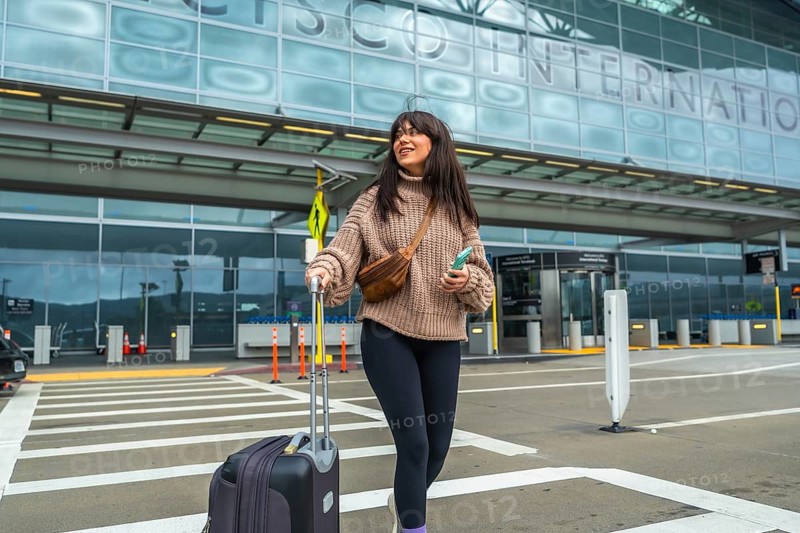 Young woman walking across a crosswalk outside a modern airport terminal, pulling luggage and checking her mobile phone as she prepares for a solo trip or business flight