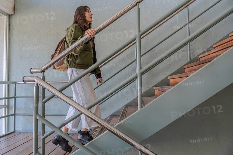 Young brunette woman in green jacket and backpack walking up interior stairs with metal railing in a modern building, looking upward with purpose
