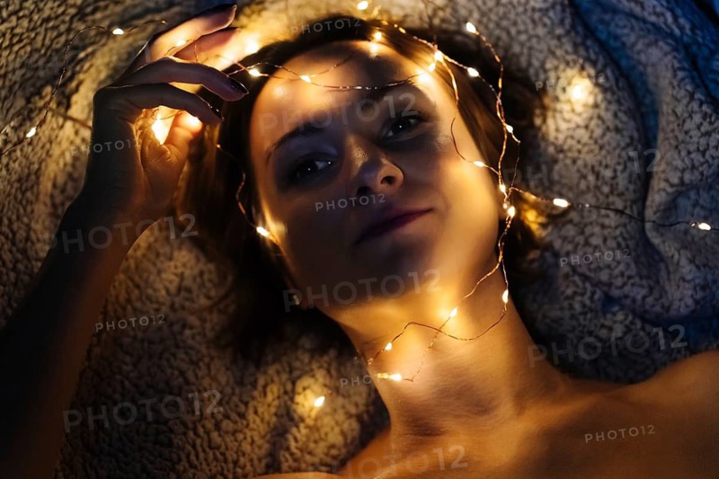 Close-up of woman's face with festive lights. Woman lies on textured blanket with warm yellow fairy lights wrapped around her head, neck and hand. Glowing lights cast soft highlights on her face and fingers, while the surrounding darkness deepens the contrast between illuminated areas and shadowed fabric. Her relaxed pose and the close framing emphasize the interplay of warm light, skin tone and textile texture, creating an intimate atmospheric portrait with pronounced depth and gentle gradients