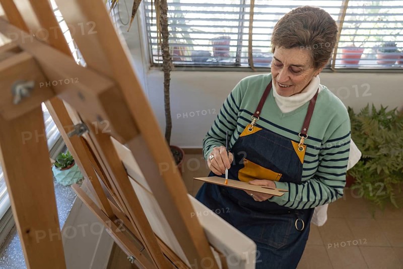 High angle view of a mature female artist smiling as she mixes paint on a small wooden palette with a fine brush, wearing a denim apron in her home art studio