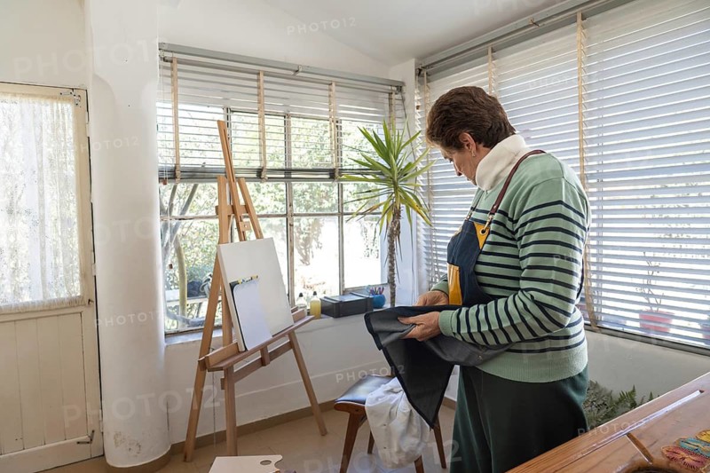 Older woman in striped sweater examines or folds a dark denim apron while standing near a wooden easel and white canvas in her sunlit home studio
