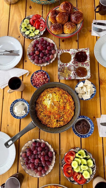 Wooden table laden with a colorful and healthy mediterranean style breakfast, featuring a large omelet in a pan, fresh grapes, sliced vegetables, sweet buns, cheese, and various spreads