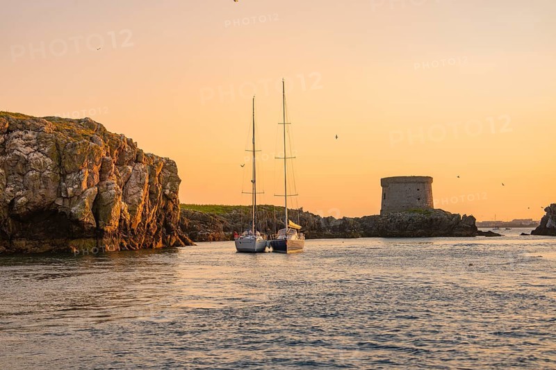 A sailboat sails between rocks near a historic tower at sunset, Howth, Ireland