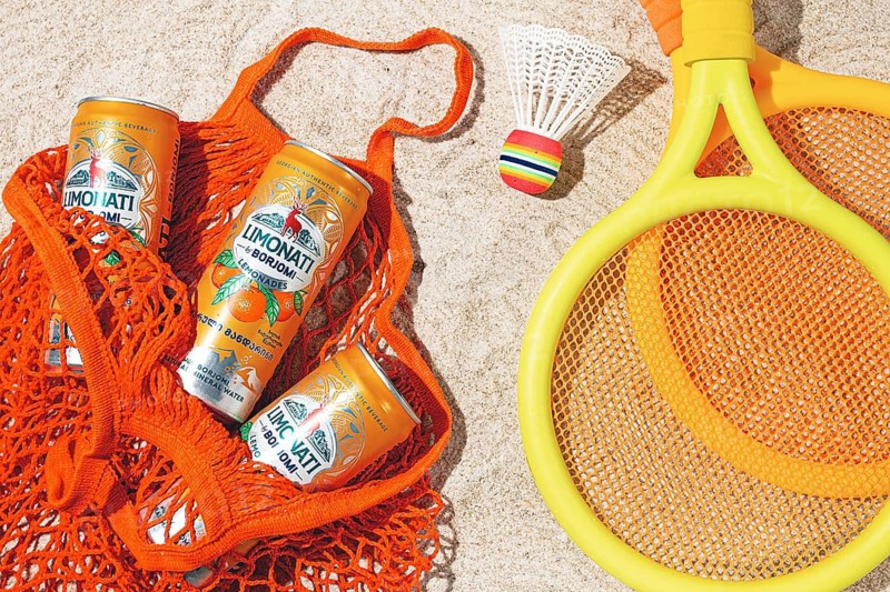 In a mesh bag, on a sandy beach, there are cans of tangerine lemonade and Borjomi drink. There are rackets and shuttlecock nearby, summer entertainment and recreation, no people